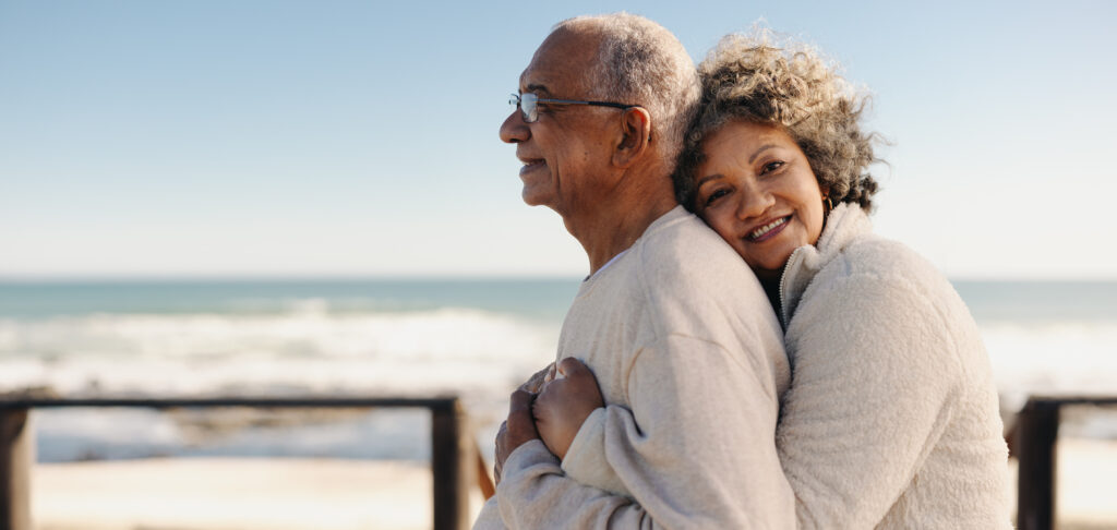 Romantic senior woman smiling at the camera while embracing her husband by the ocean. Affectionate elderly couple enjoying spending some quality time and snowbird planning together after retirement.
