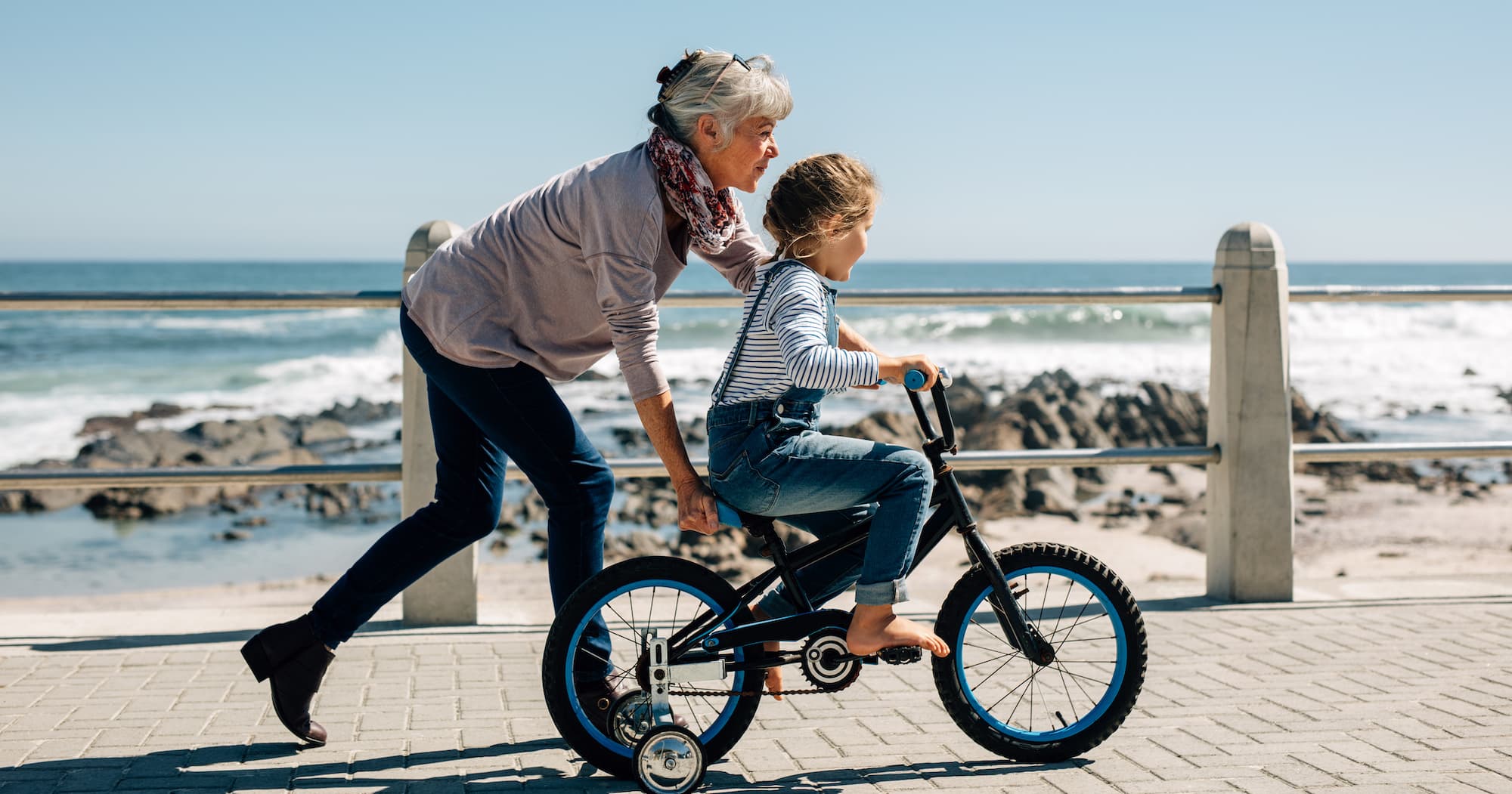 Side view of a girl riding a bicycle while her grandmother runs along holding the kid. Senior woman teaching a small girl to ride a bicycle on the road alongside the beach.